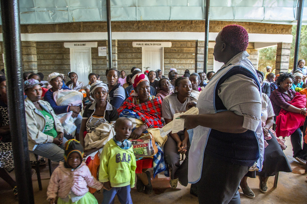 A health worker addressing a seated community gathering outdoors.