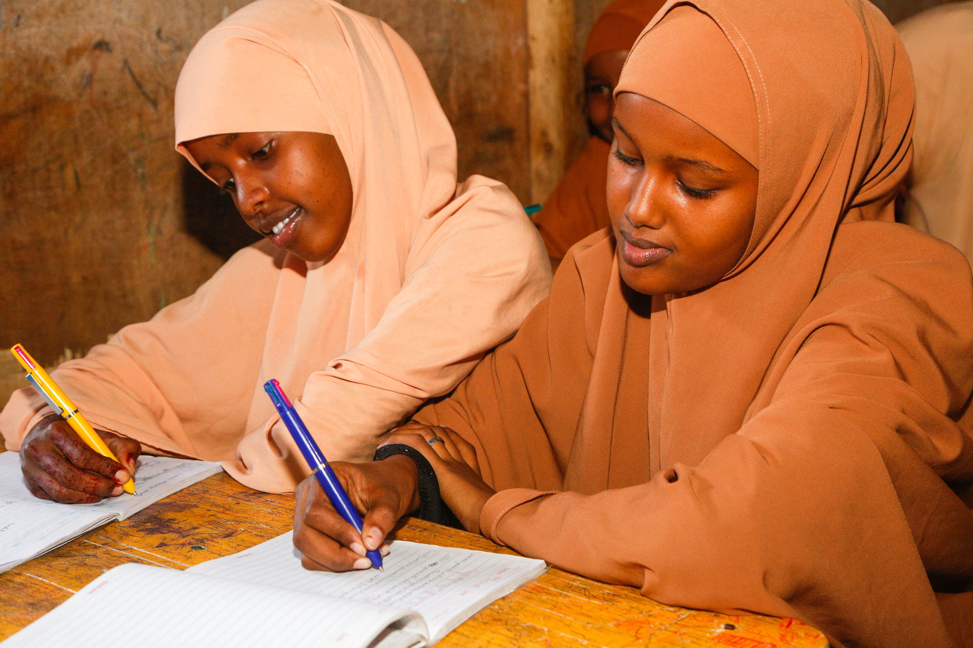 Two students writing in exercise books together at a desk in warm light.